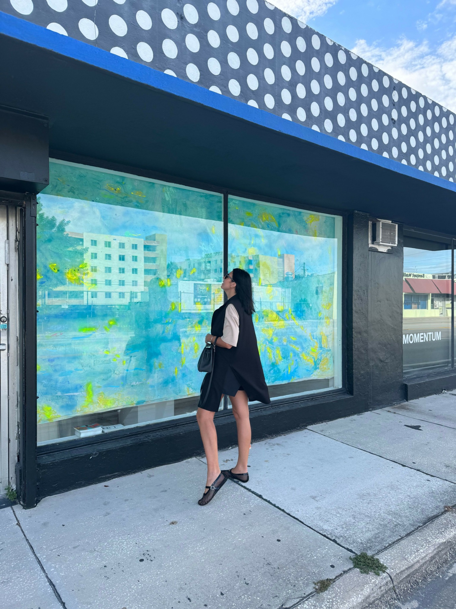 Woman standing outside a building with large colorful window display, wearing a black dress and heels, holding a bag.