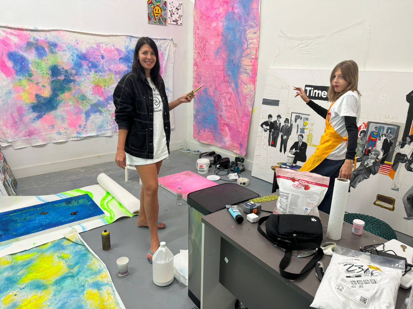 Two women in an art studio with colorful paintings and supplies, one pointing at a pink and blue vertical artwork.