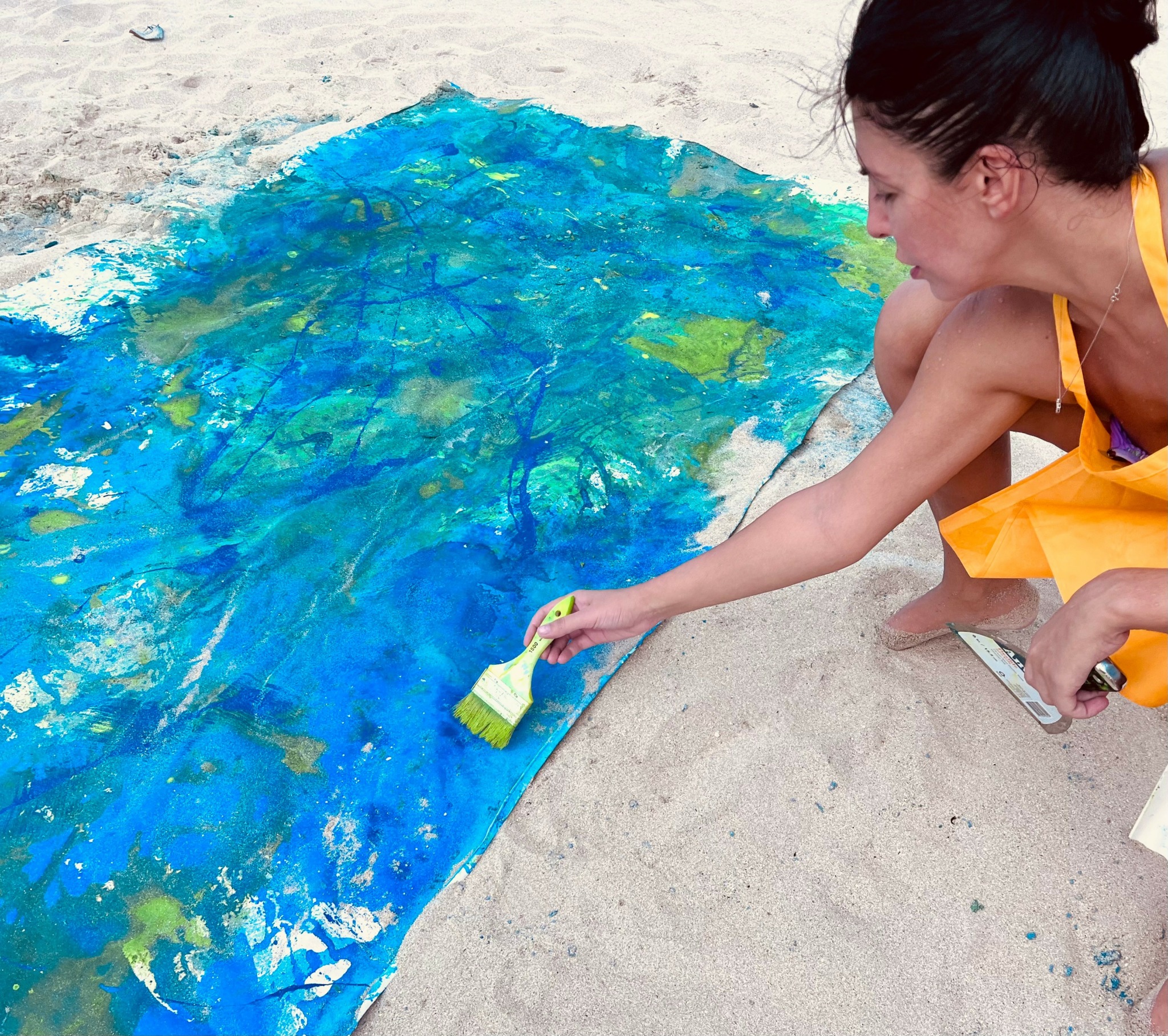 Woman painting a large blue and green abstract artwork on the sand with a brush.