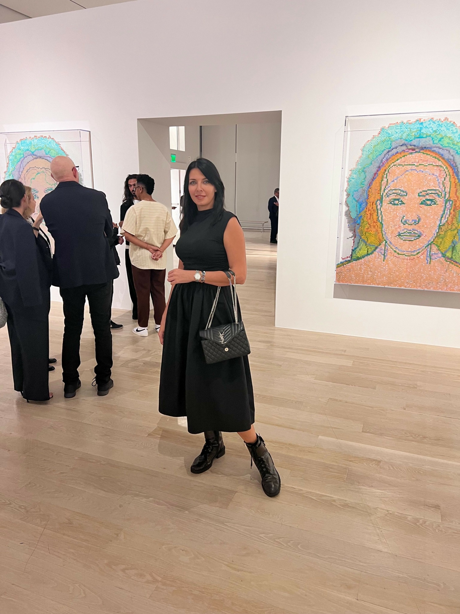 Woman in black dress holding a bag, standing in art gallery with colorful portraits on white walls.