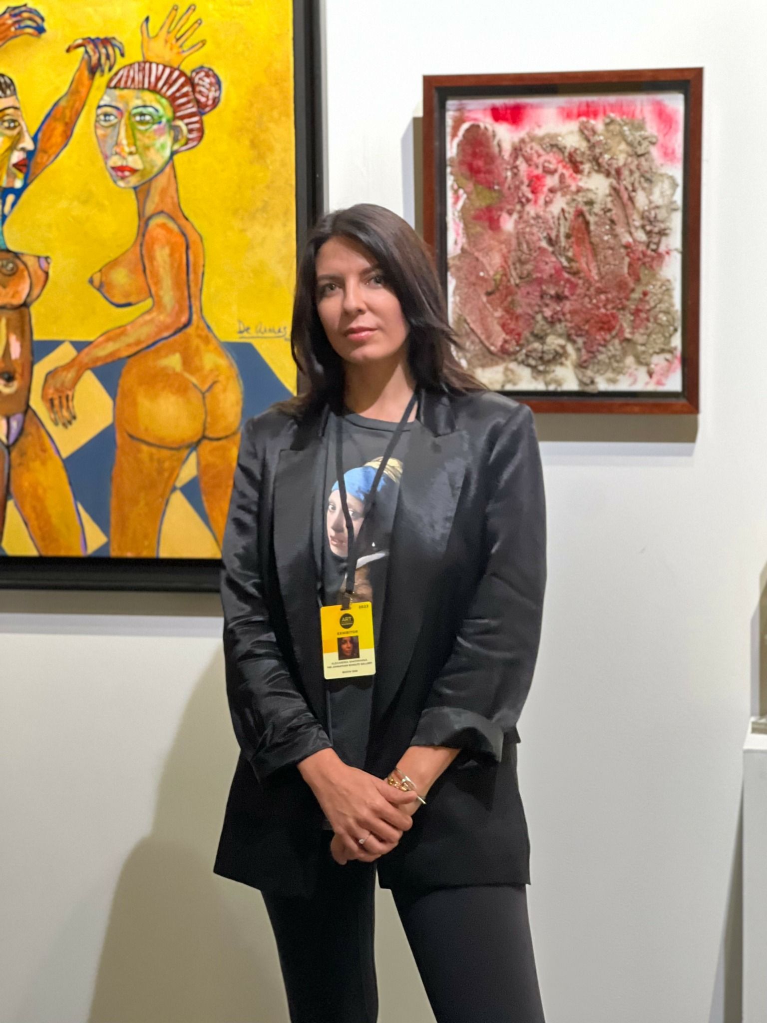 Woman with dark hair wearing a black jacket and lanyard, standing in front of colorful artwork in an art gallery.