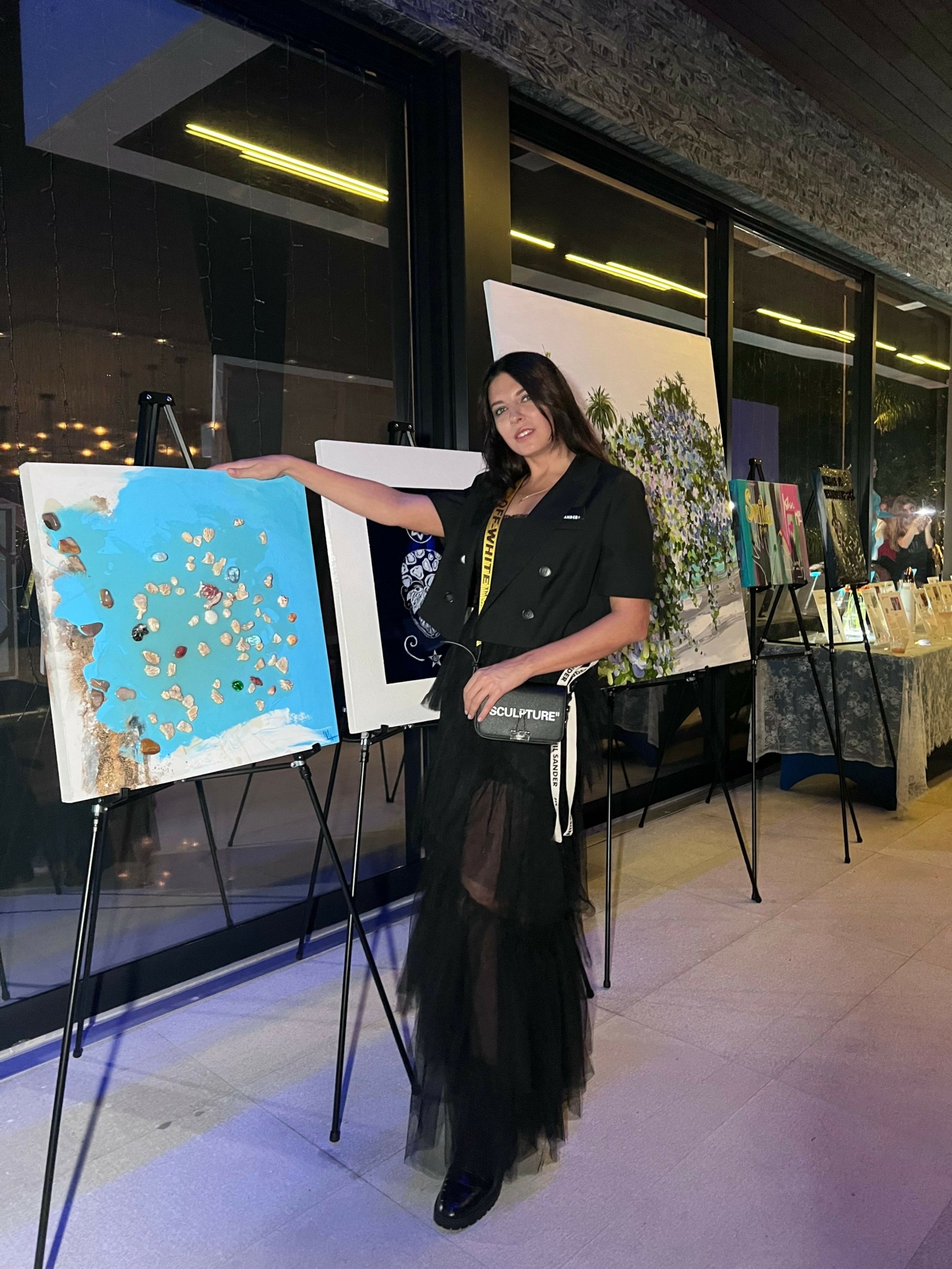 Woman in black dress stands next to paintings on display in an indoor gallery.