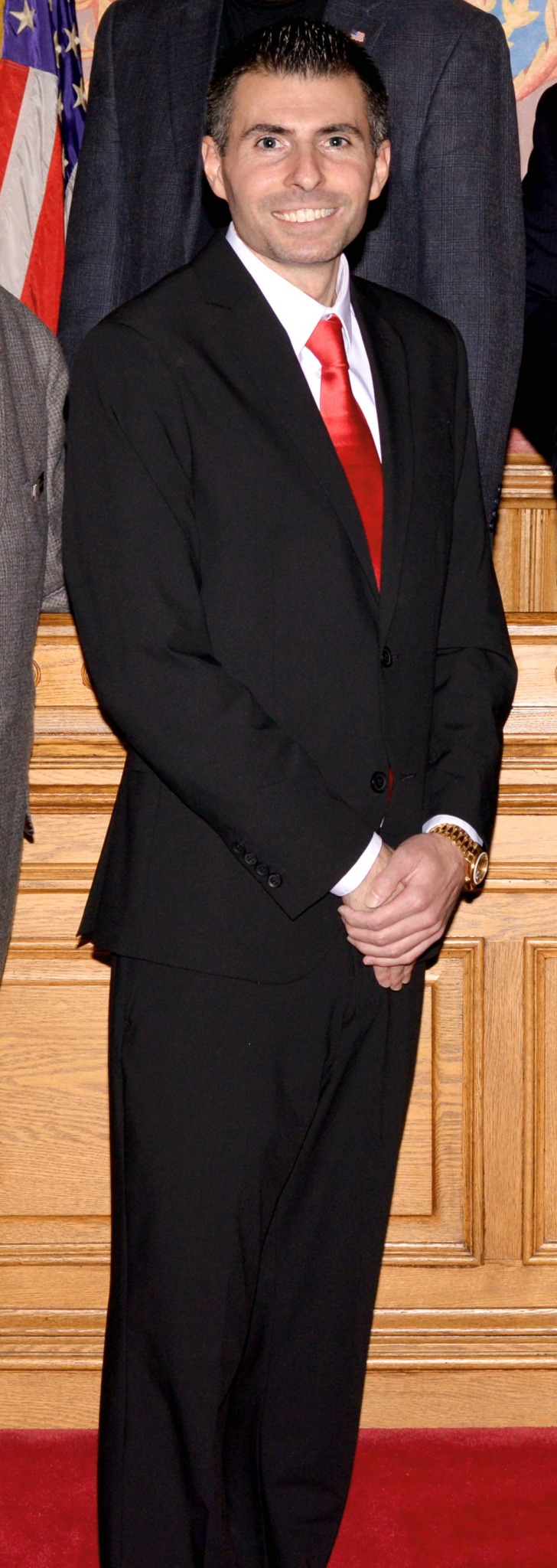 Man in black suit with red tie standing indoors with wooden paneling behind him.