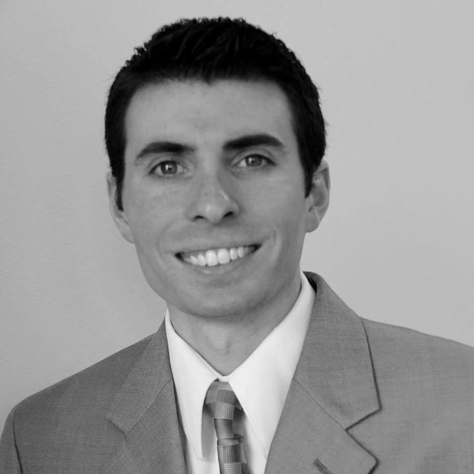 Man with dark hair in a suit and tie, smiling, facing forward.