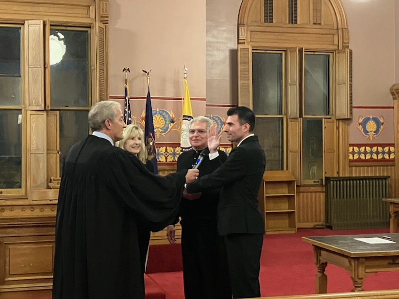 Three men in formal attire shaking hands in a room with wooden paneling and flags in the background.