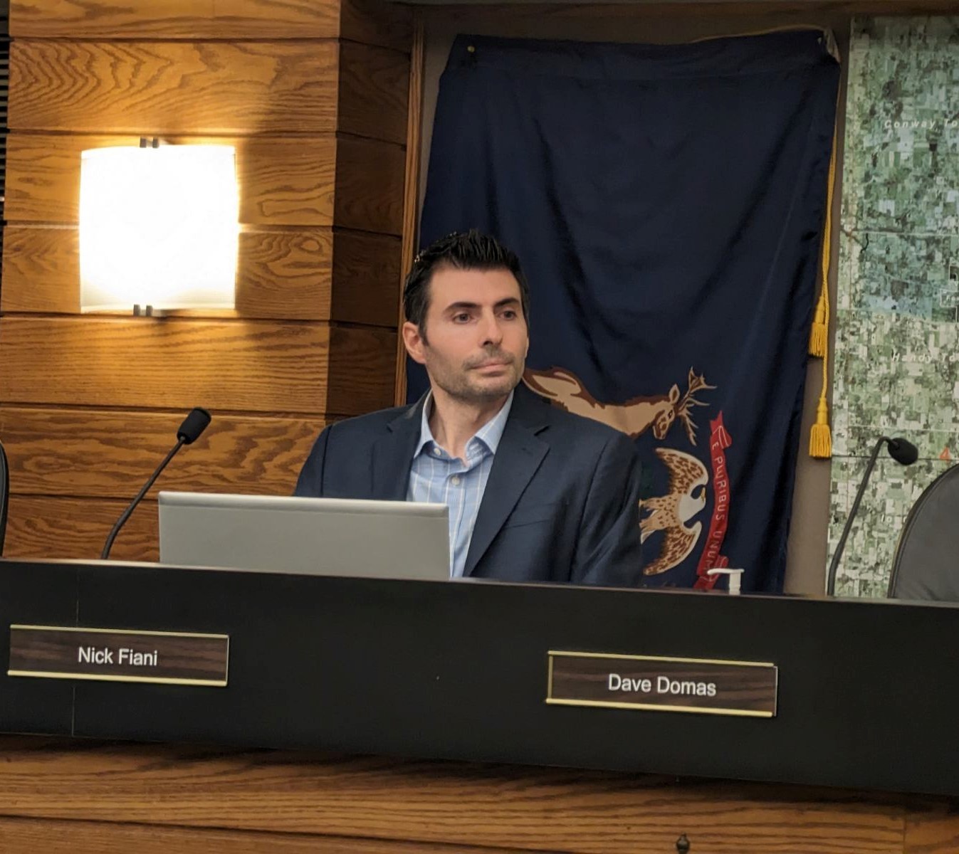 Man in suit sitting at a desk with a laptop, in a room with wood paneling and a dark curtain.
