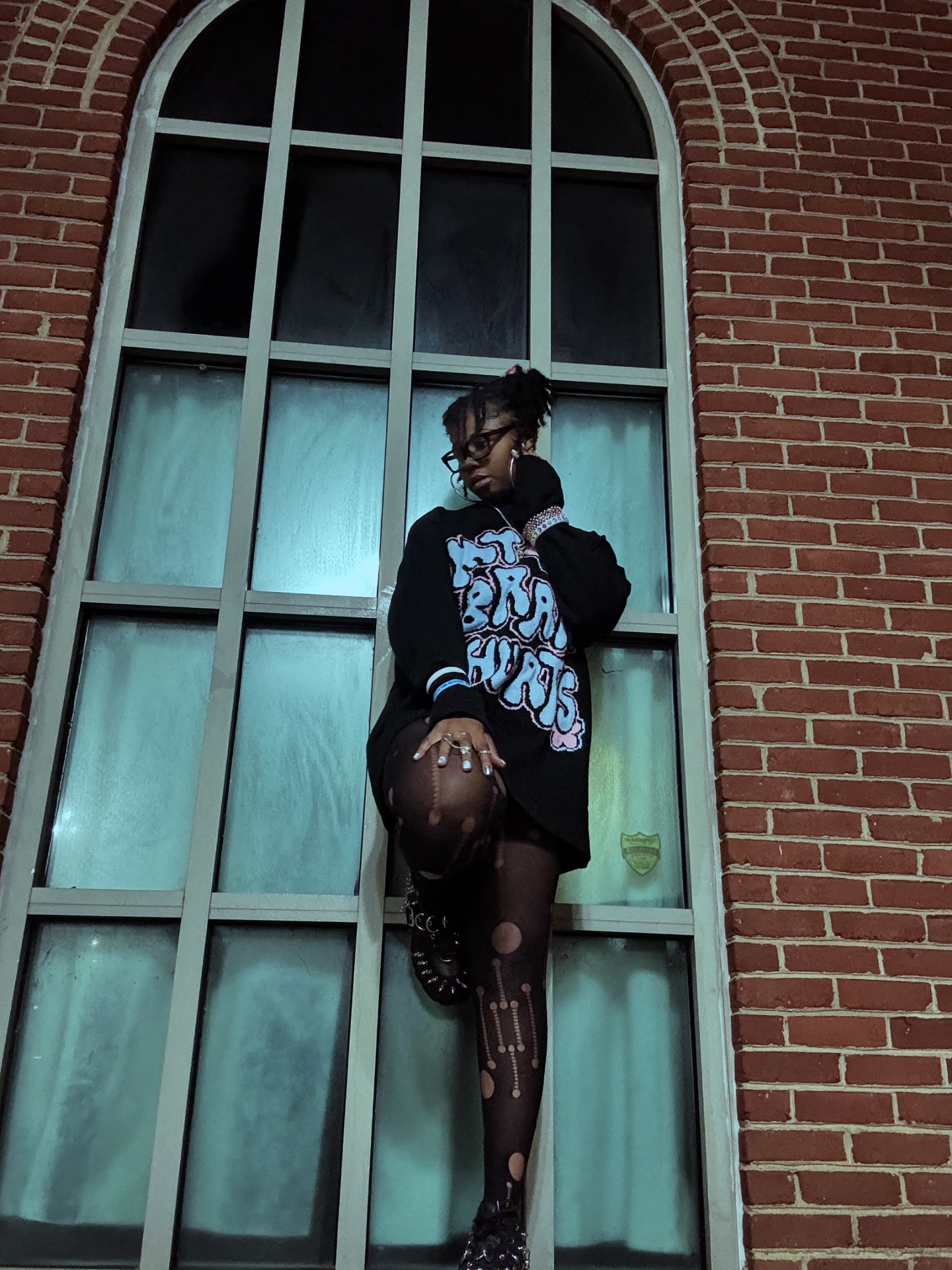 Person standing in front of large arched window with brick wall, holding a basketball, wearing a black jacket and patterned tights.