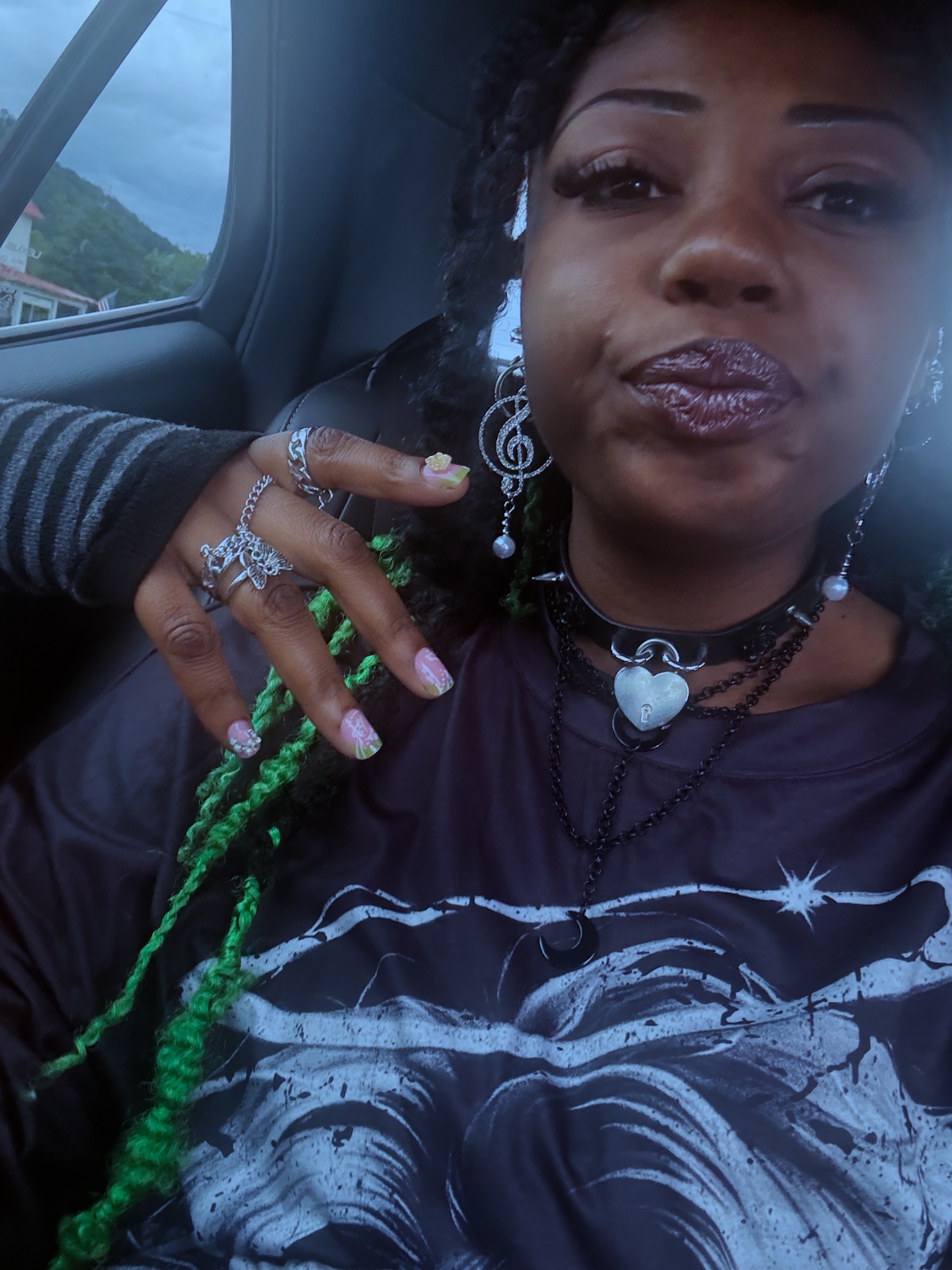 Close-up of a woman with dark hair and jewelry, sitting inside a vehicle, with a window showing a landscape outside.