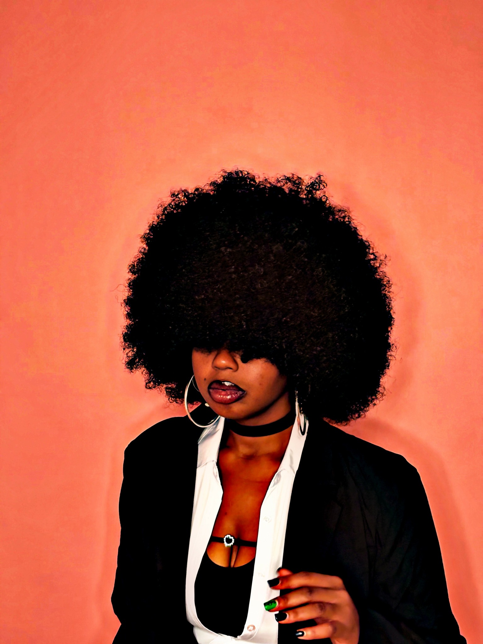 Woman with large curly afro hairstyle, dark lipstick, wearing a black jacket and white shirt, against pink background.