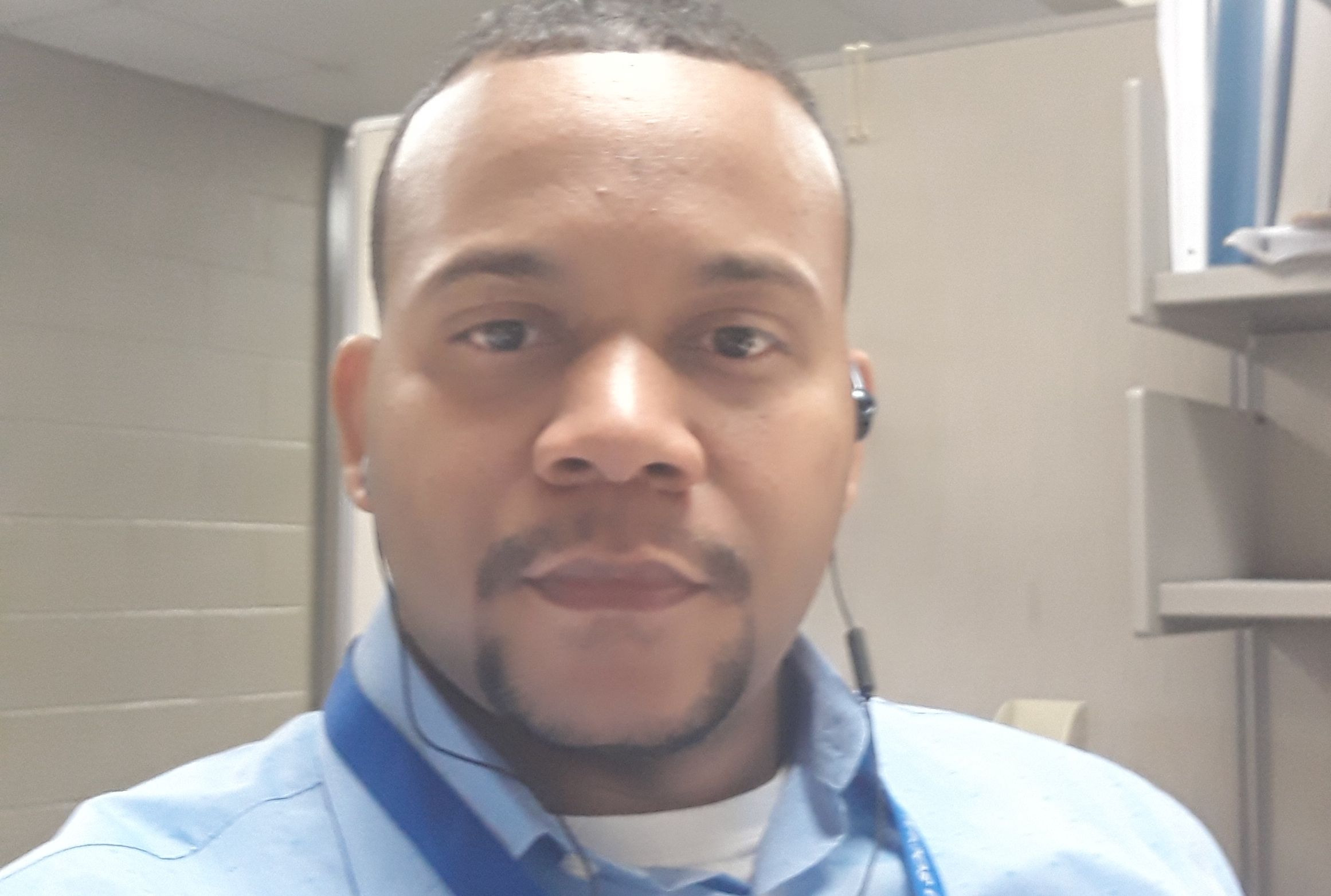 Close-up of a man with short hair and a goatee, wearing a blue shirt and earphones, in an indoor setting.