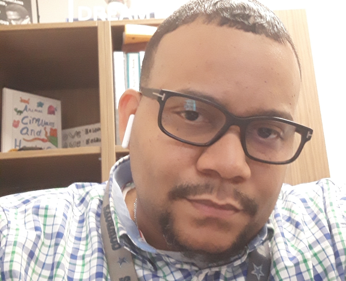 Man with glasses and earbuds, wearing a checkered shirt, in an indoor setting with bookshelves in the background.