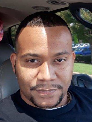 Close-up of a man with short hair and a slight smile inside a vehicle, sunlight partially illuminating his face.