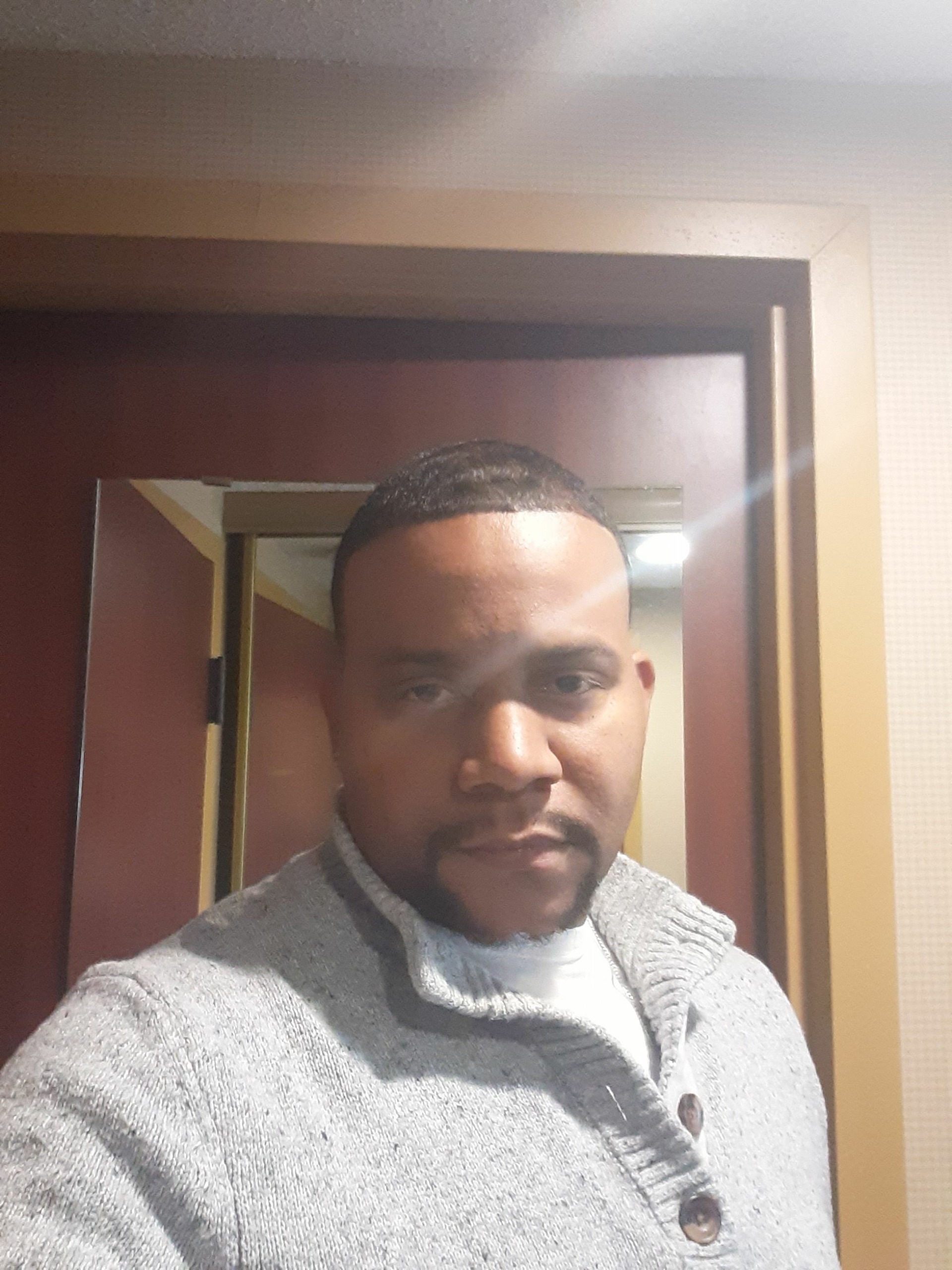 Portrait of a man with short hair and a beard, wearing a gray sweater, standing indoors near a doorframe.