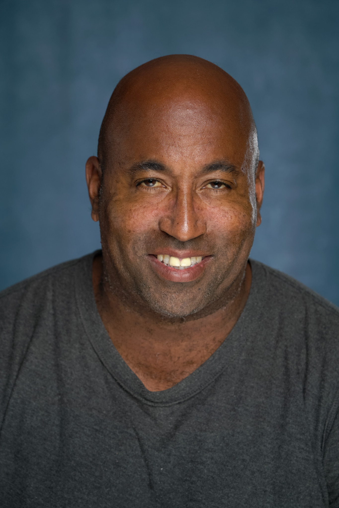 Smiling man with a shaved head and dark shirt against a blue background.