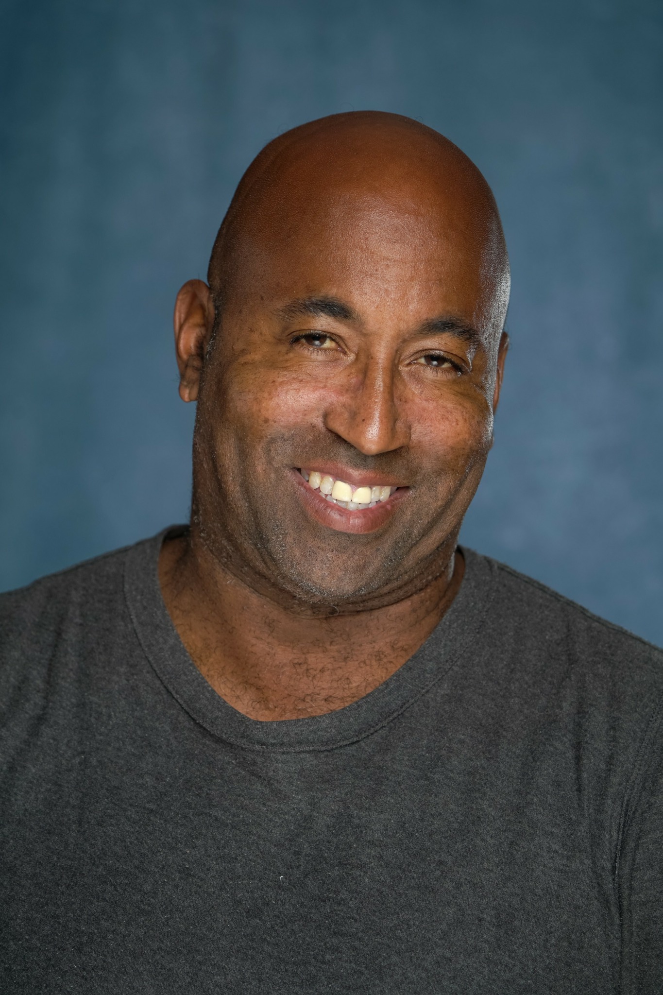 Man with a bald head smiling, wearing a dark gray shirt, against a blue background.