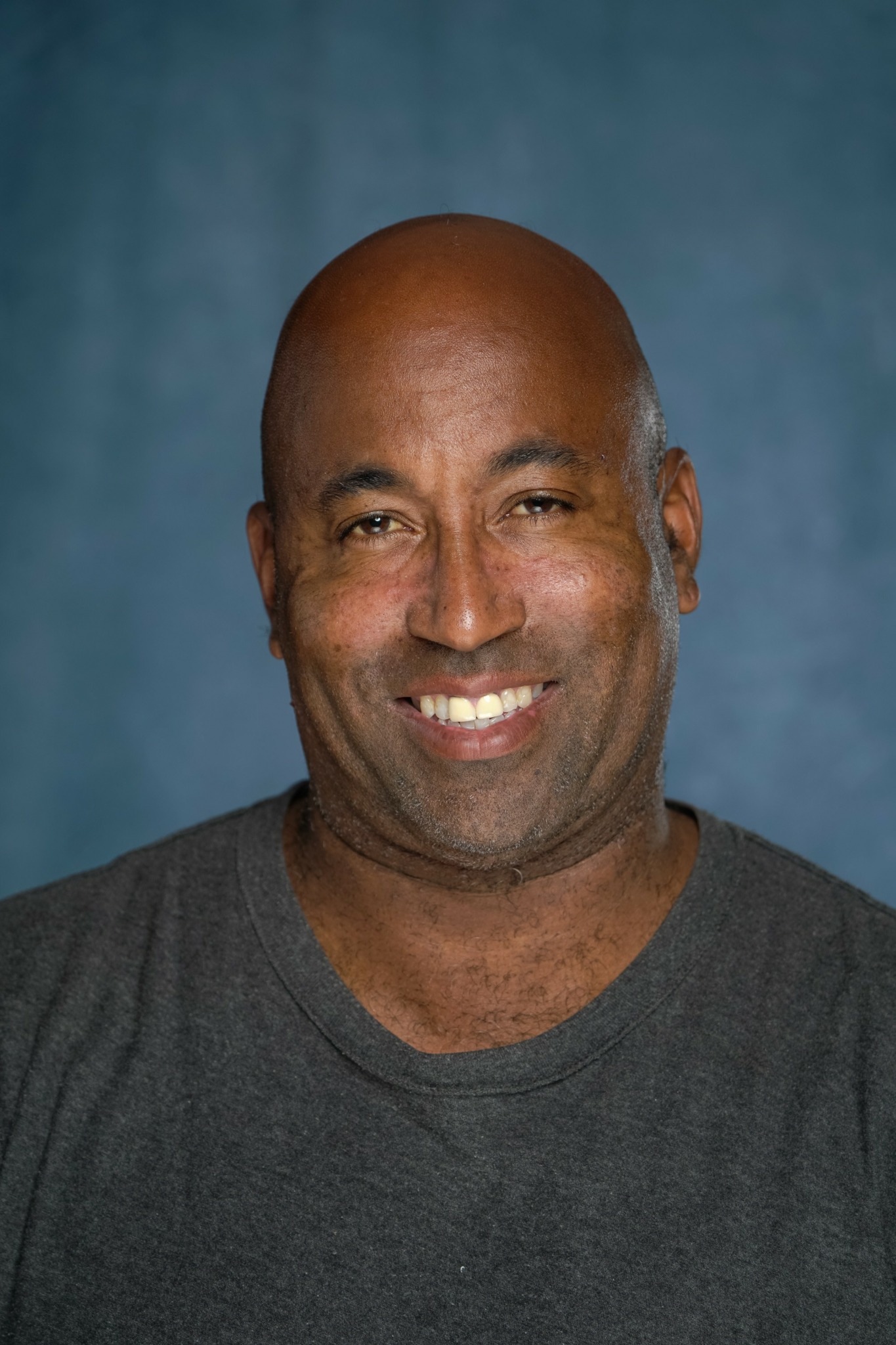 Smiling man with a bald head and dark shirt against a blue background.