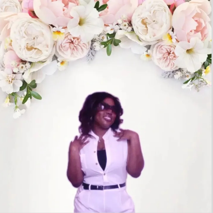 Person with dark hair and sunglasses smiling, wearing a white sleeveless shirt and pants, standing in front of floral background.