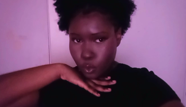 Person with short curly hair making a pose with hand near chin, looking at camera, against a plain background.