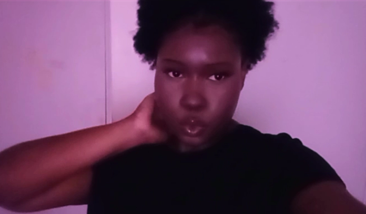 Person with short curly hair wearing a black shirt, touching neck, in front of a plain wall.