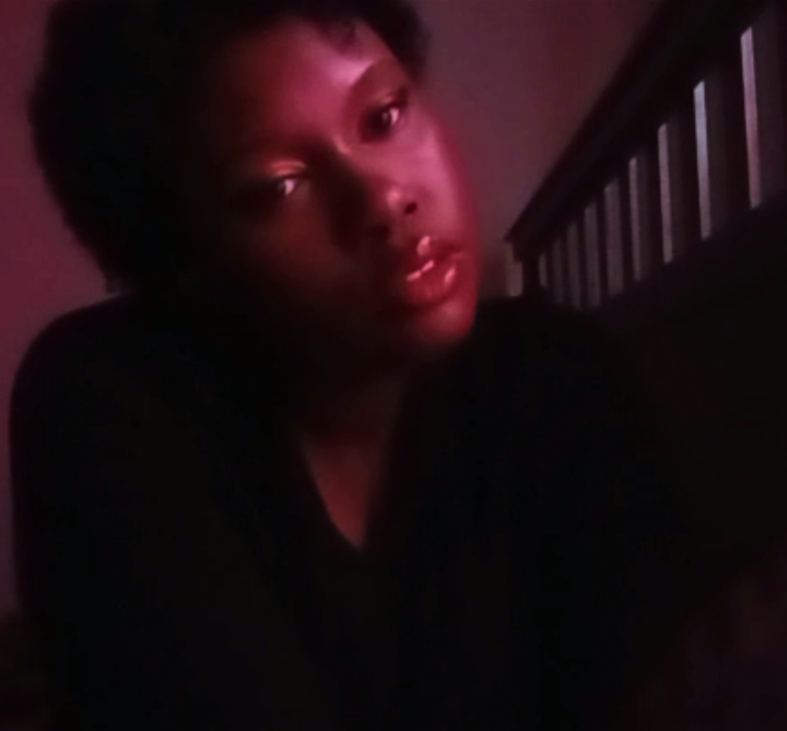 Person with short hair and dark clothing looking at camera, dimly lit background with a railing.