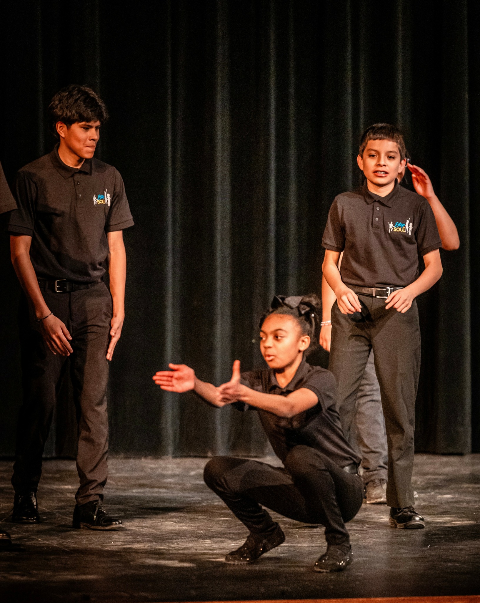 Three young performers on stage, one crouching and gesturing, two standing, in front of a dark curtain.