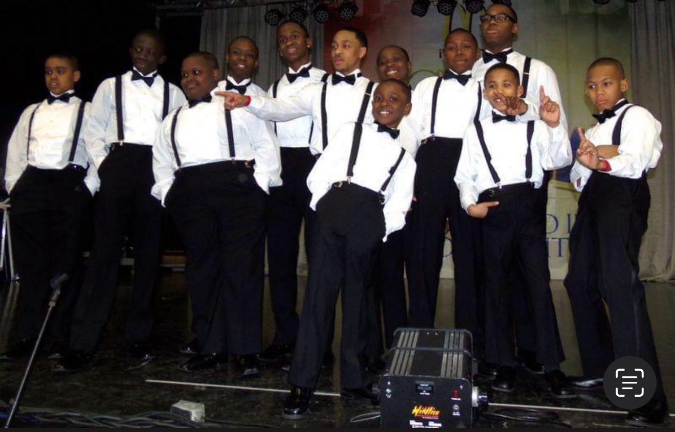 Group of children dressed in formal attire standing on stage, some with arms crossed or hands in pockets.
