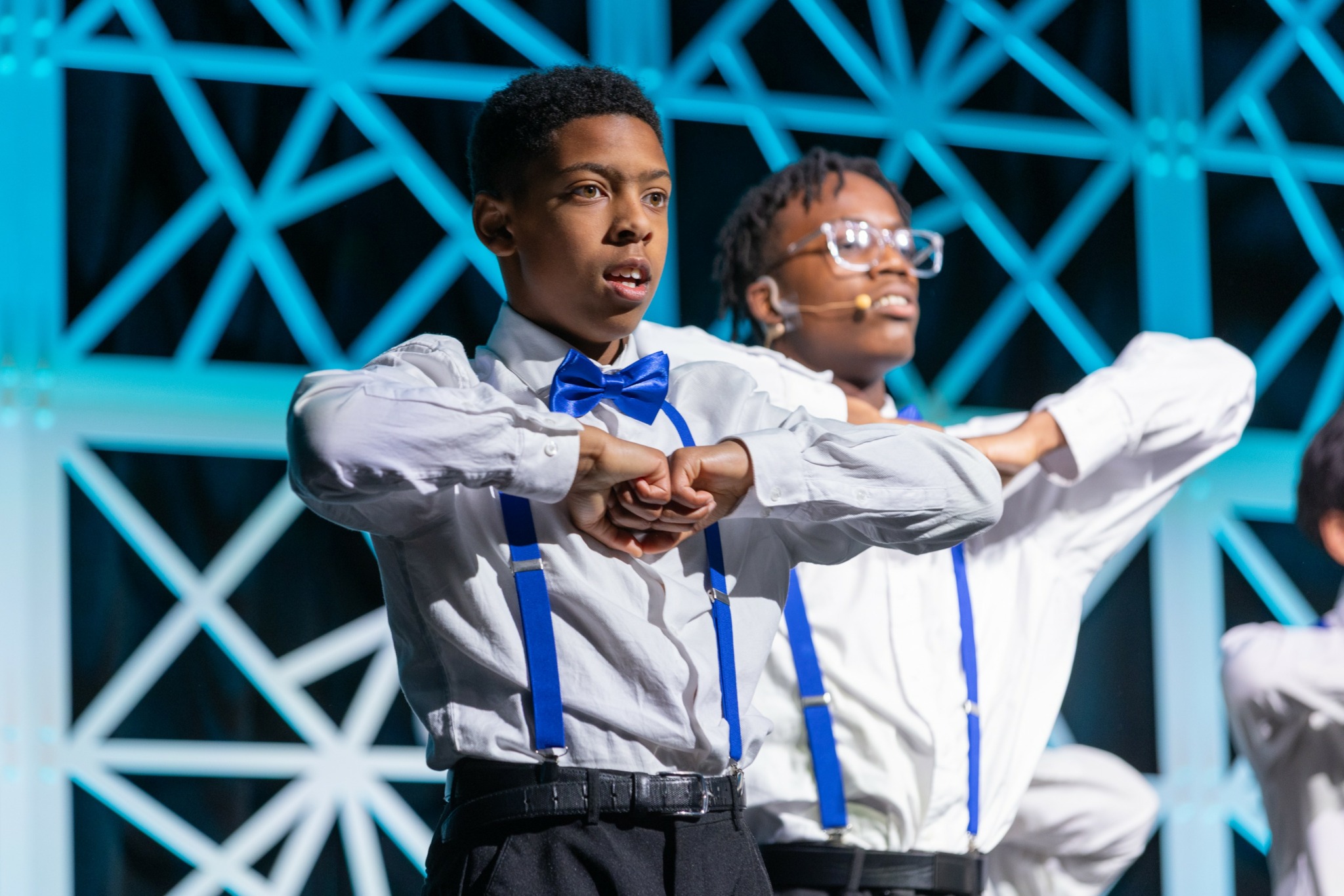 Two children in formal attire perform a synchronized dance move on stage with a geometric blue background.