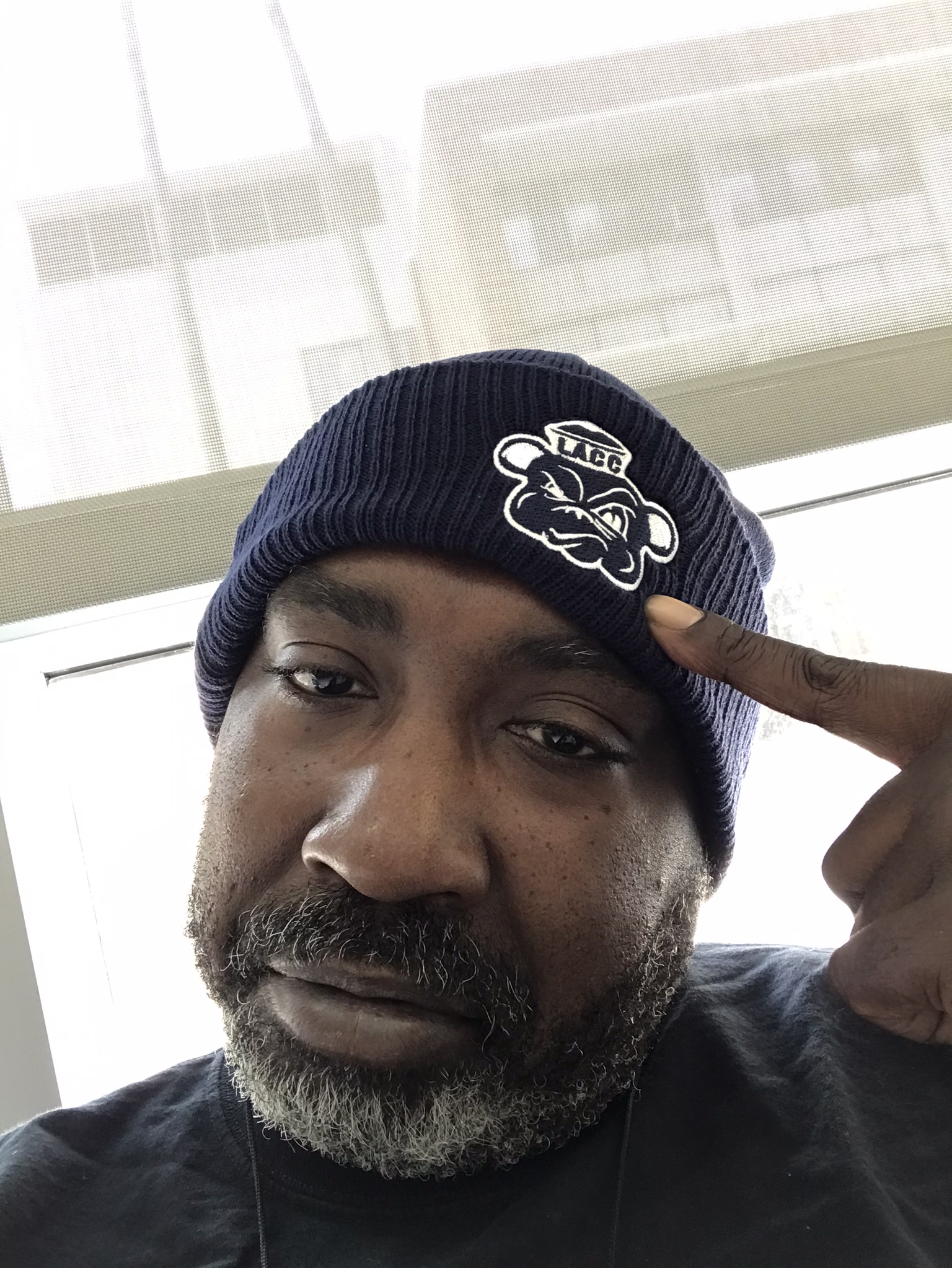 Man wearing a navy blue beanie with a bear logo, pointing to his temple, indoors with windows behind him.