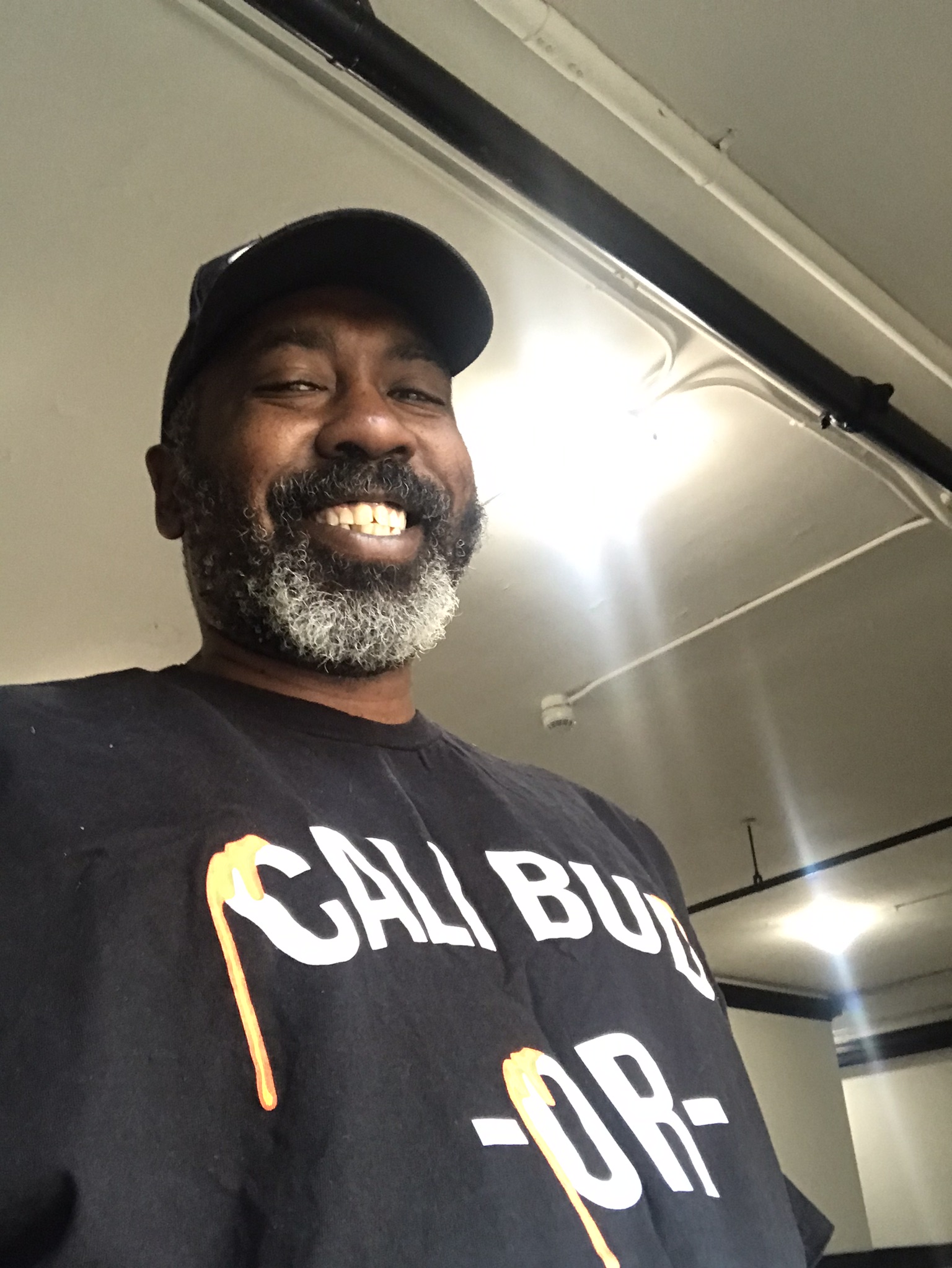 Smiling man with a beard wearing a black cap and a black T-shirt with white text.