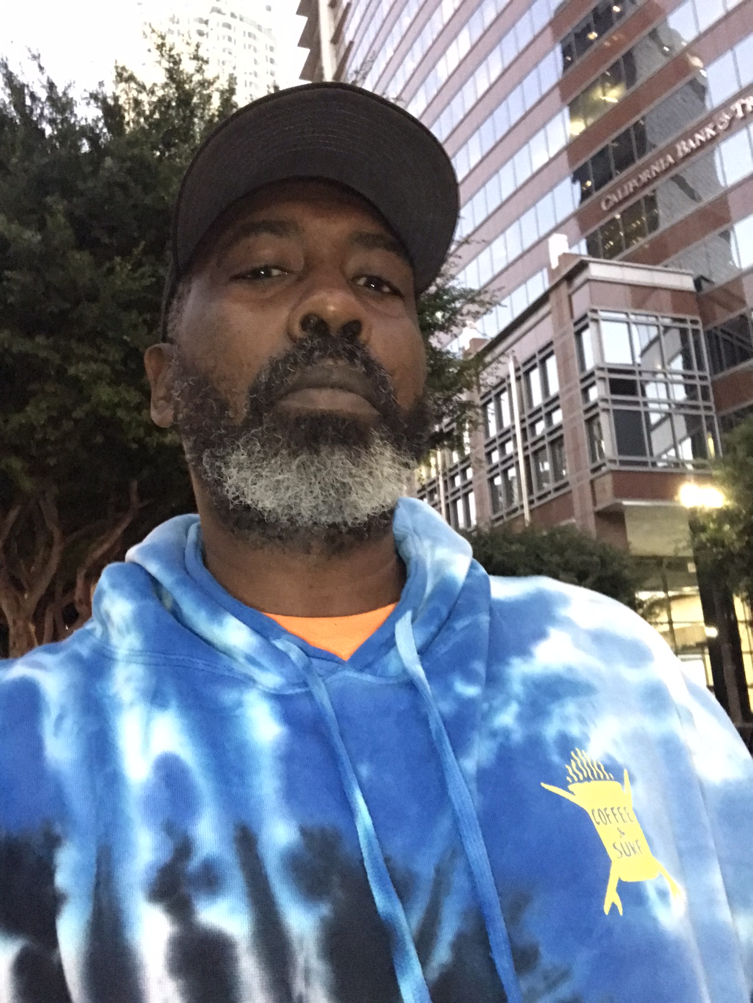 Man with a beard wearing a cap and a blue tie-dye hoodie outdoors with buildings in the background.