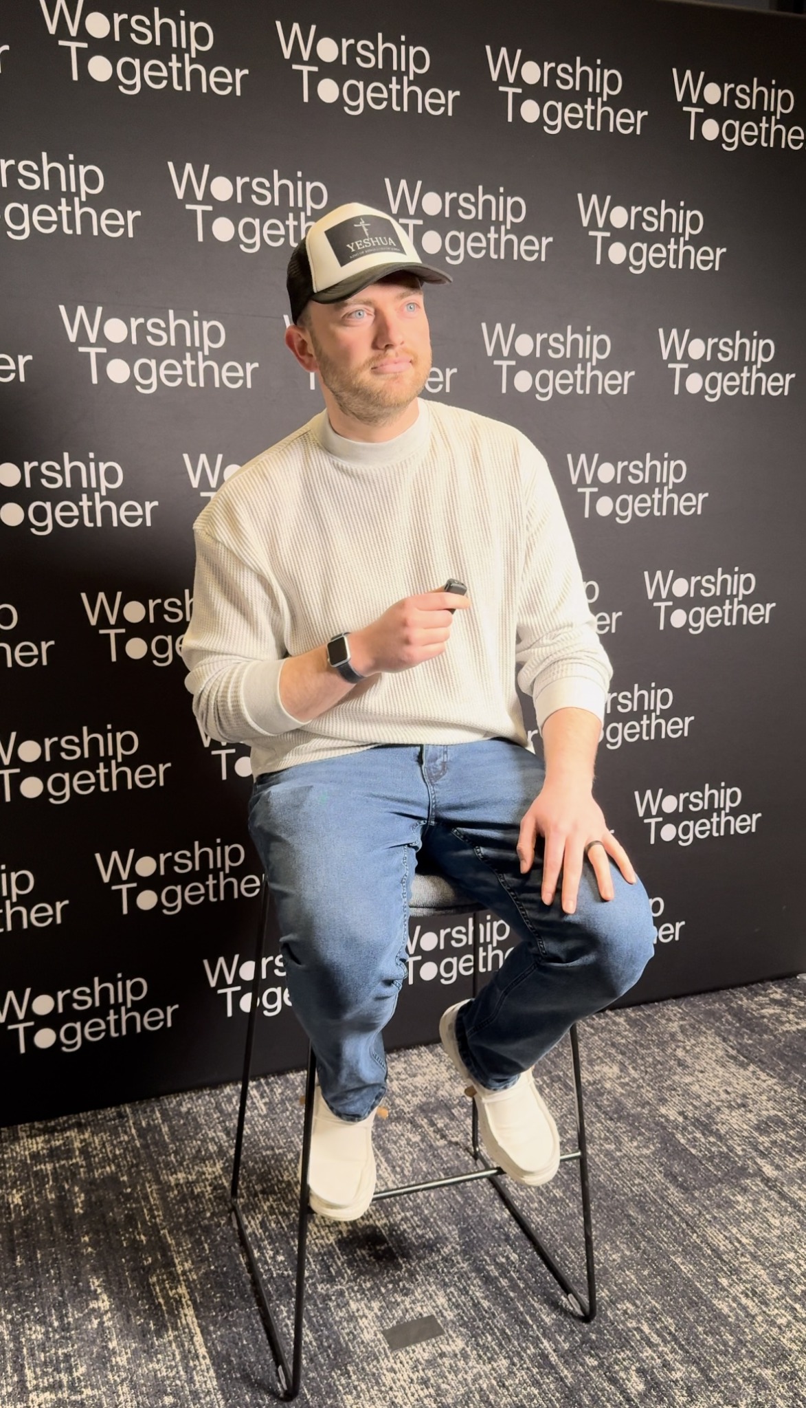 Man sitting on a stool holding a microphone, wearing a white sweater, jeans, and a cap, in front of a backdrop.