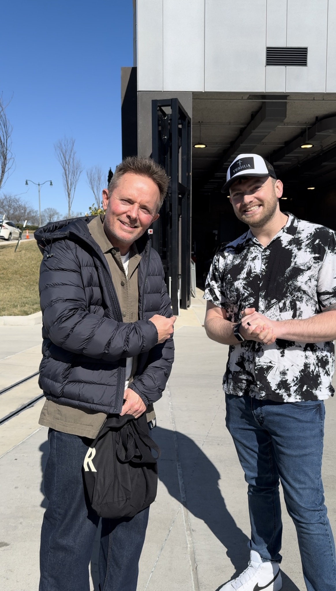 Two men shaking hands outdoors near a modern building, smiling, with clear blue sky in background.