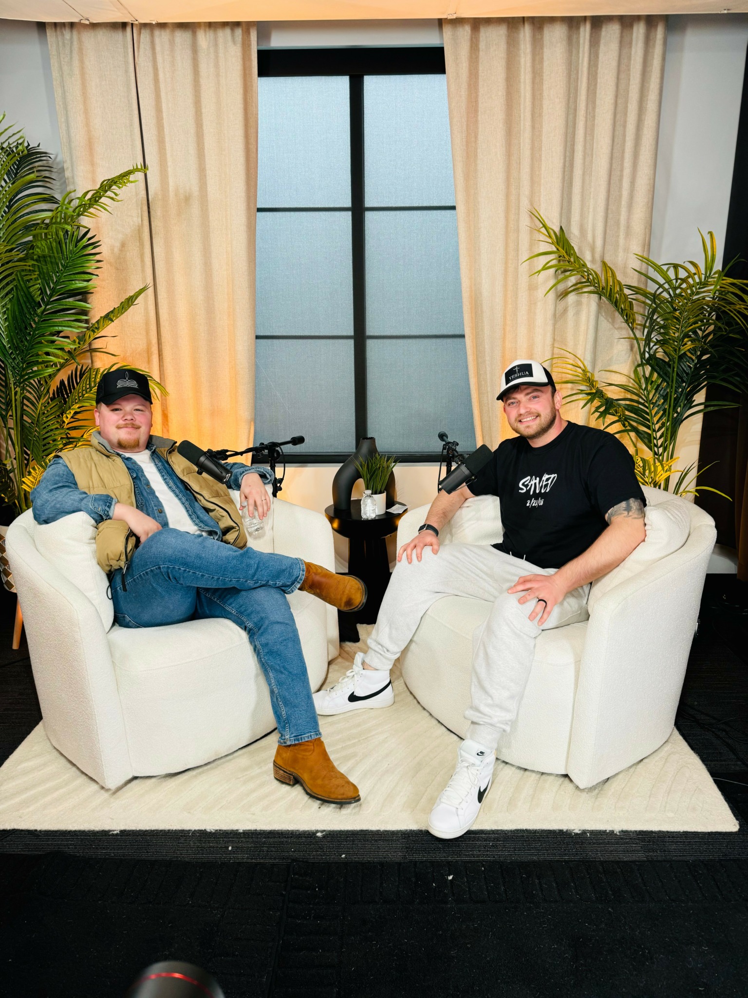 Two men sit on white sofas with plants and a window with curtains behind them, holding rifles and smiling.