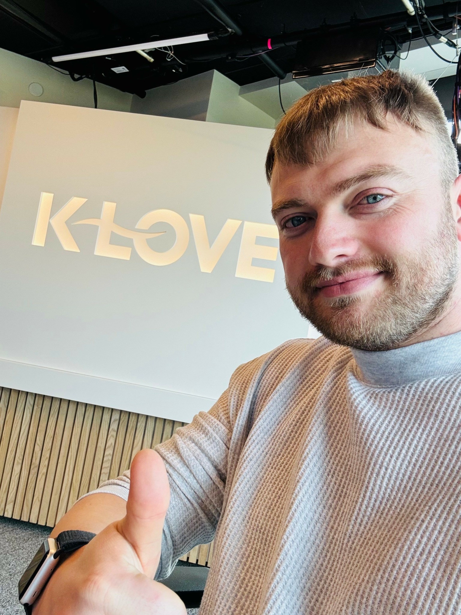 Young man with short hair and beard smiling, giving a thumbs-up in front of a white wall with a logo.
