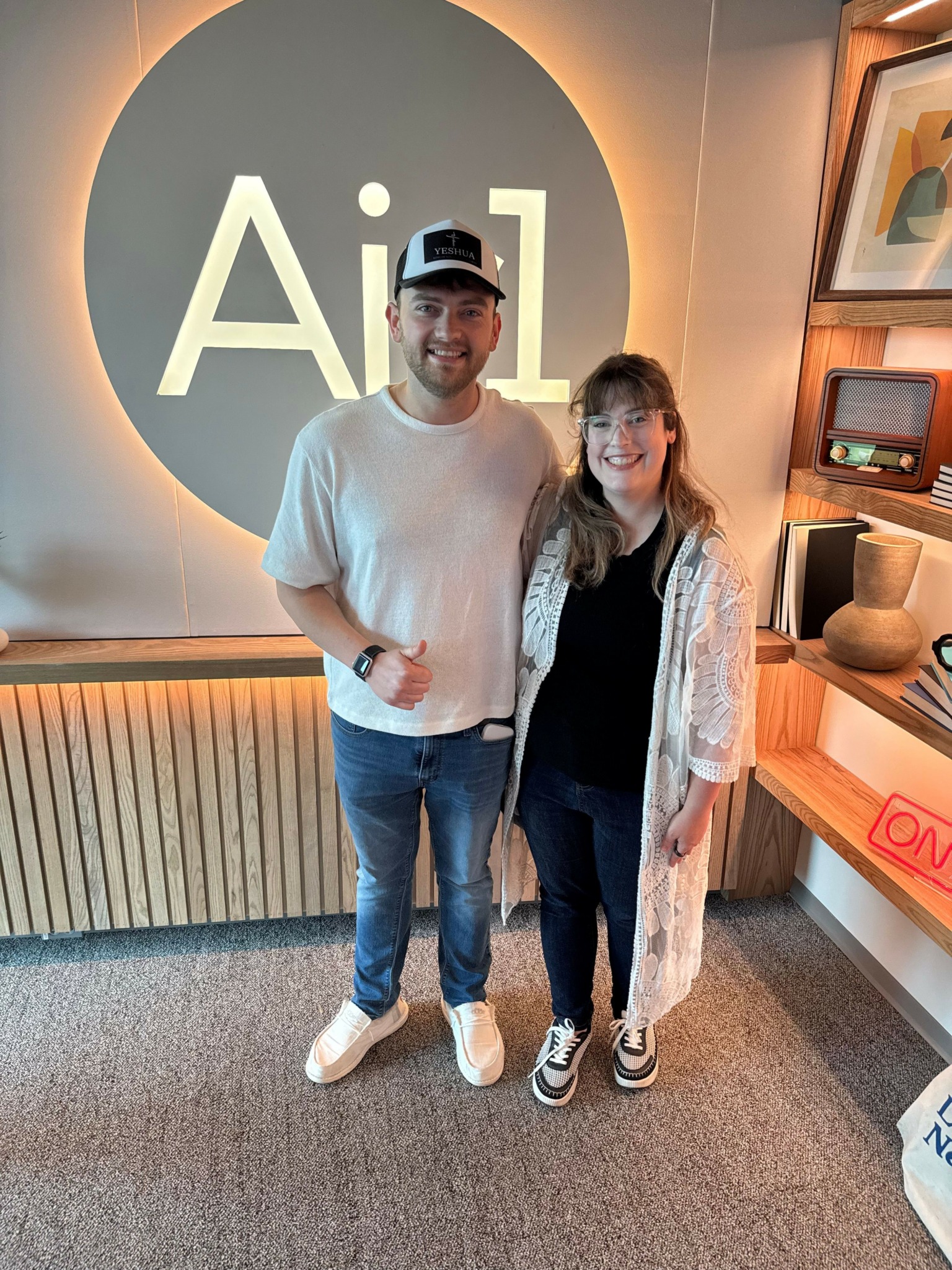 Two people standing indoors in front of a large circular sign with 'AiL' text, smiling at the camera.