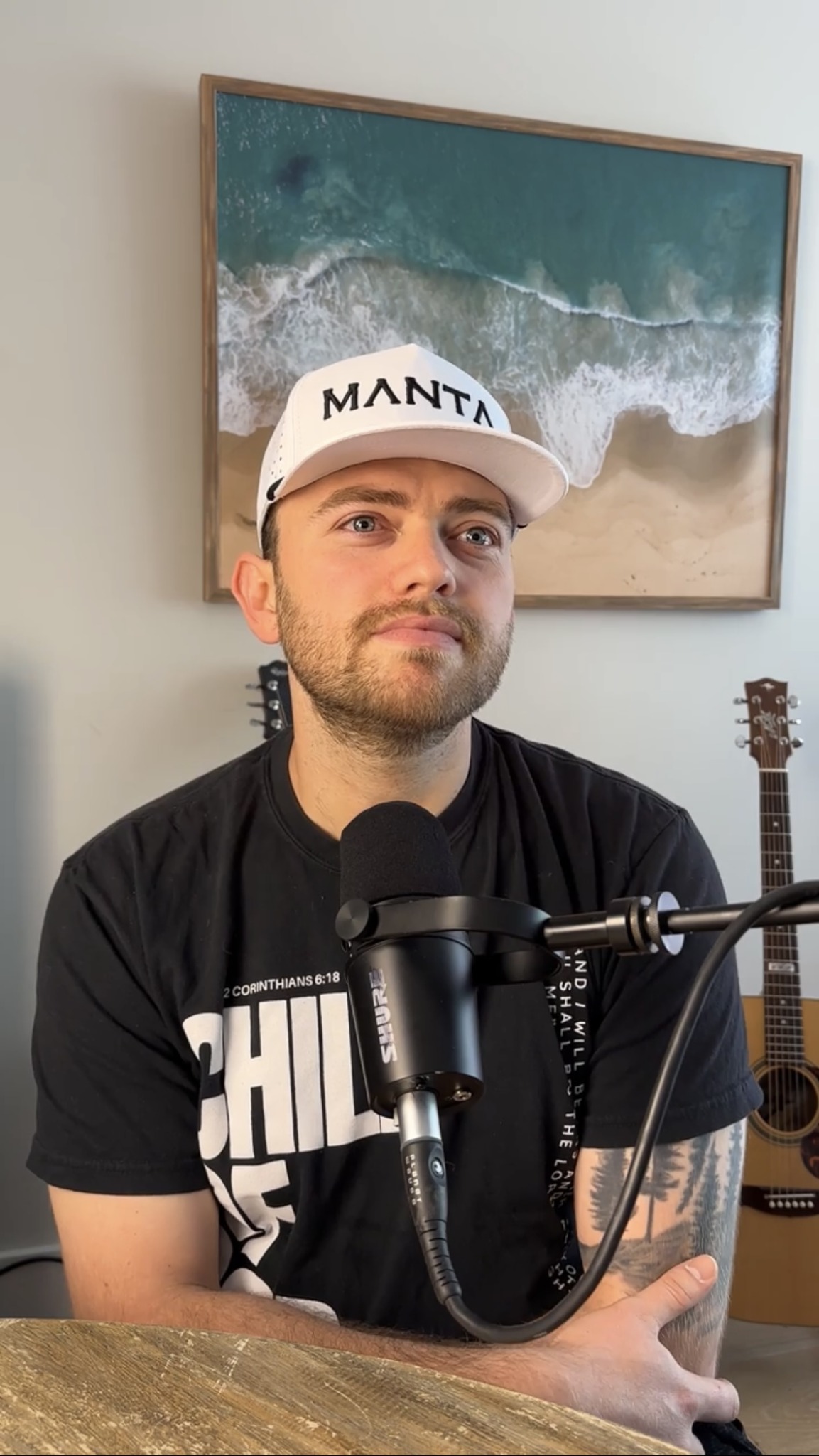 Young man wearing a white cap and black T-shirt sitting at a table with a microphone, in front of a beach photo.