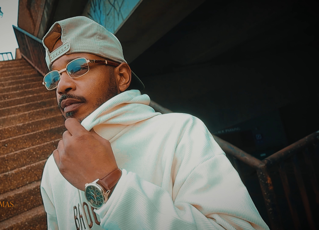 Man wearing sunglasses, a cap, and a white hoodie, standing near stairs with a thoughtful pose.