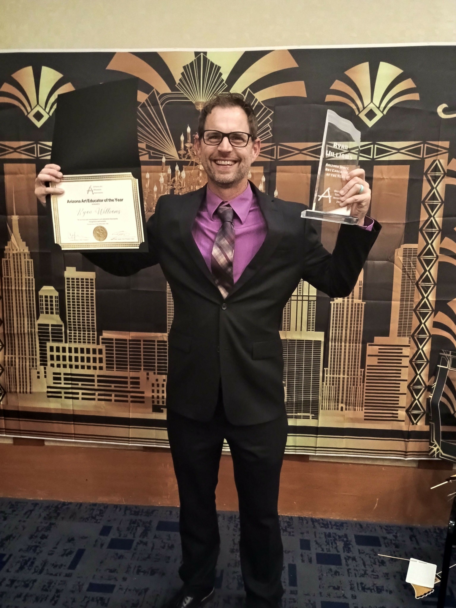 Man in suit holding a certificate and an award, smiling, standing in front of an art deco cityscape backdrop.