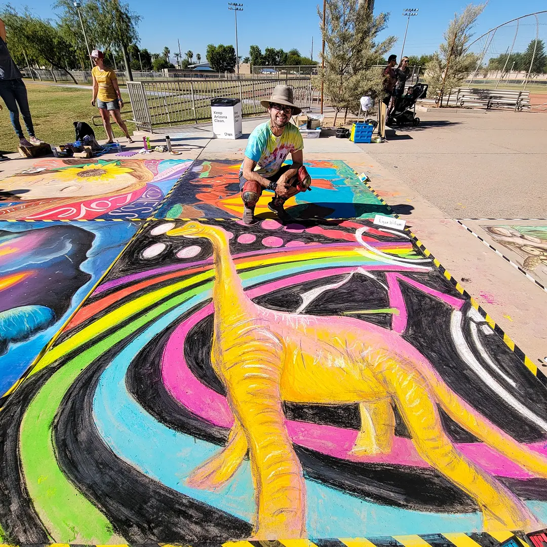 Person crouching next to colorful dinosaur chalk art on pavement at outdoor park.