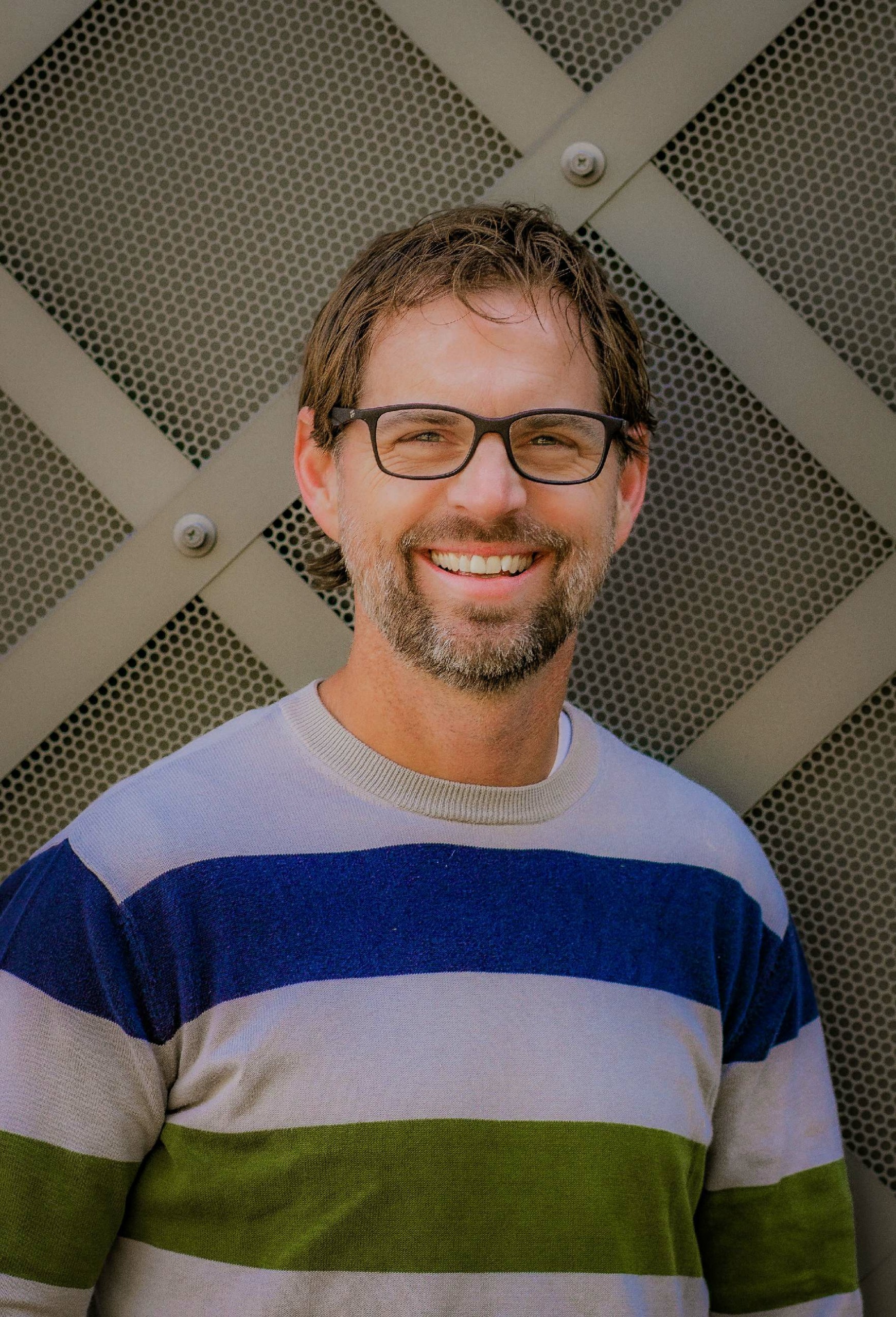 Smiling man with glasses and beard wearing a striped sweater standing outdoors.