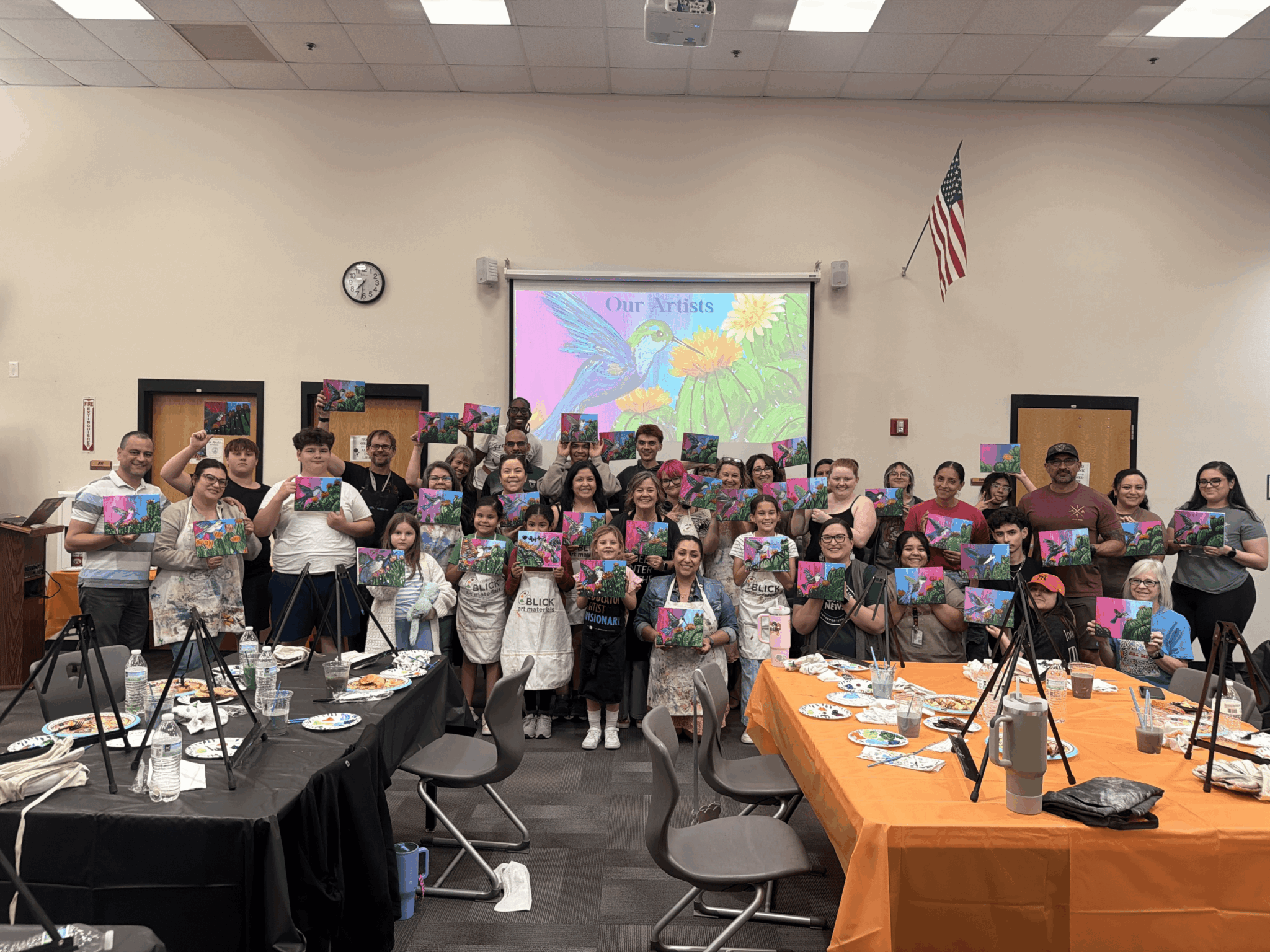 Group of children and adults in a classroom holding colorful artwork, with a screen displaying a butterfly and flowers behind them.