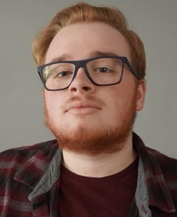 Young man with glasses and reddish hair, wearing a plaid shirt and maroon t-shirt, looking at the camera.