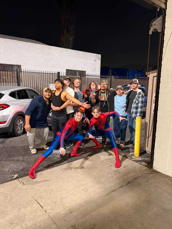 Group of people standing outdoors at night, two individuals in Spider-Man costumes crouching in front.