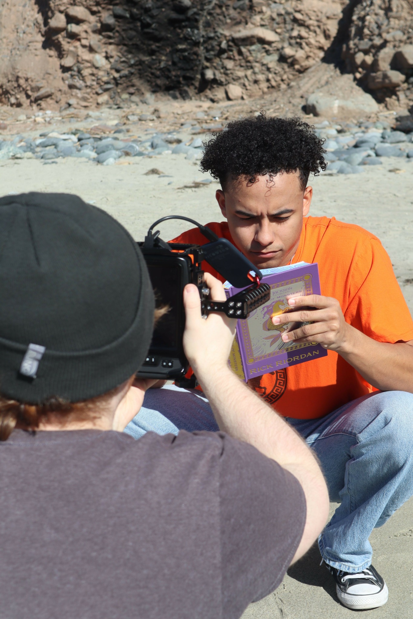 Young man with curly hair in orange shirt sitting outdoors, looking at a card, with a person filming him.