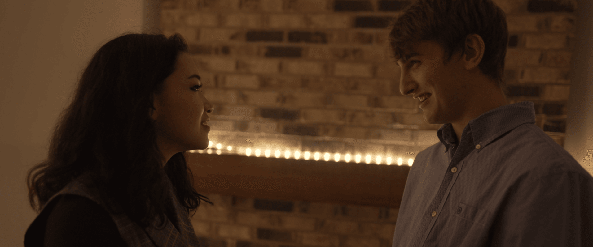 Two people facing each other indoors with a brick wall background and warm lighting.