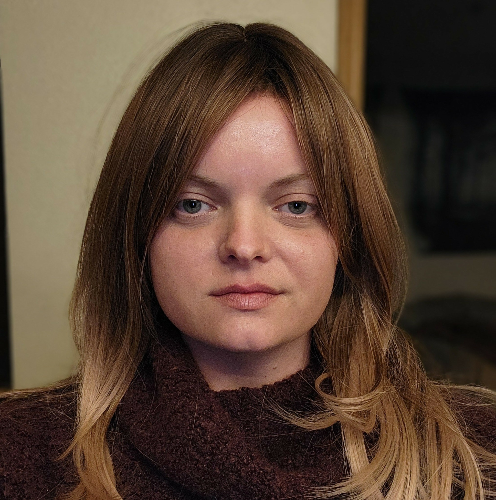 Young woman with long, wavy brown hair and blue eyes, wearing a dark turtleneck, looking directly at the camera.