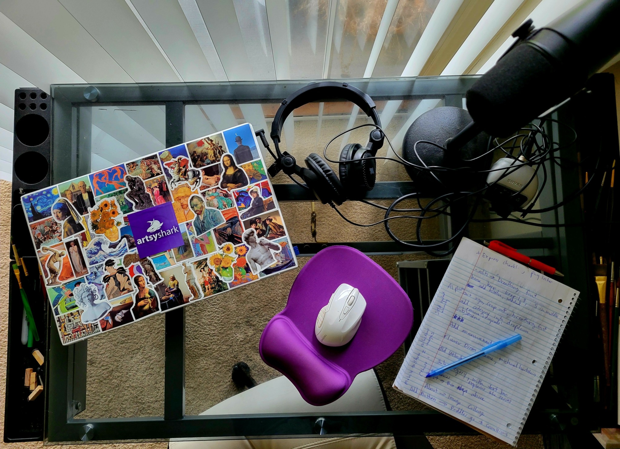 Glass desk with collage, headphones, microphone, purple mouse pad with mouse, and notebook with pen.
