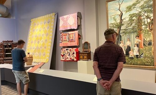 Two men in an art gallery, one looking at a display and the other looking out the window, with artwork on the wall.