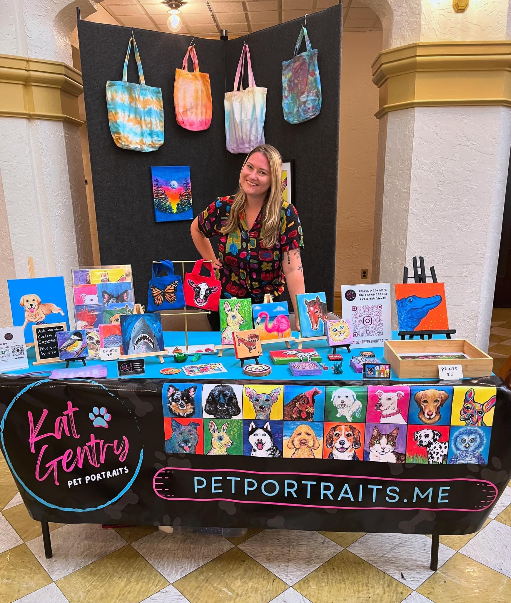 Woman standing behind a pet portrait display table with colorful artwork and tote bags in background.