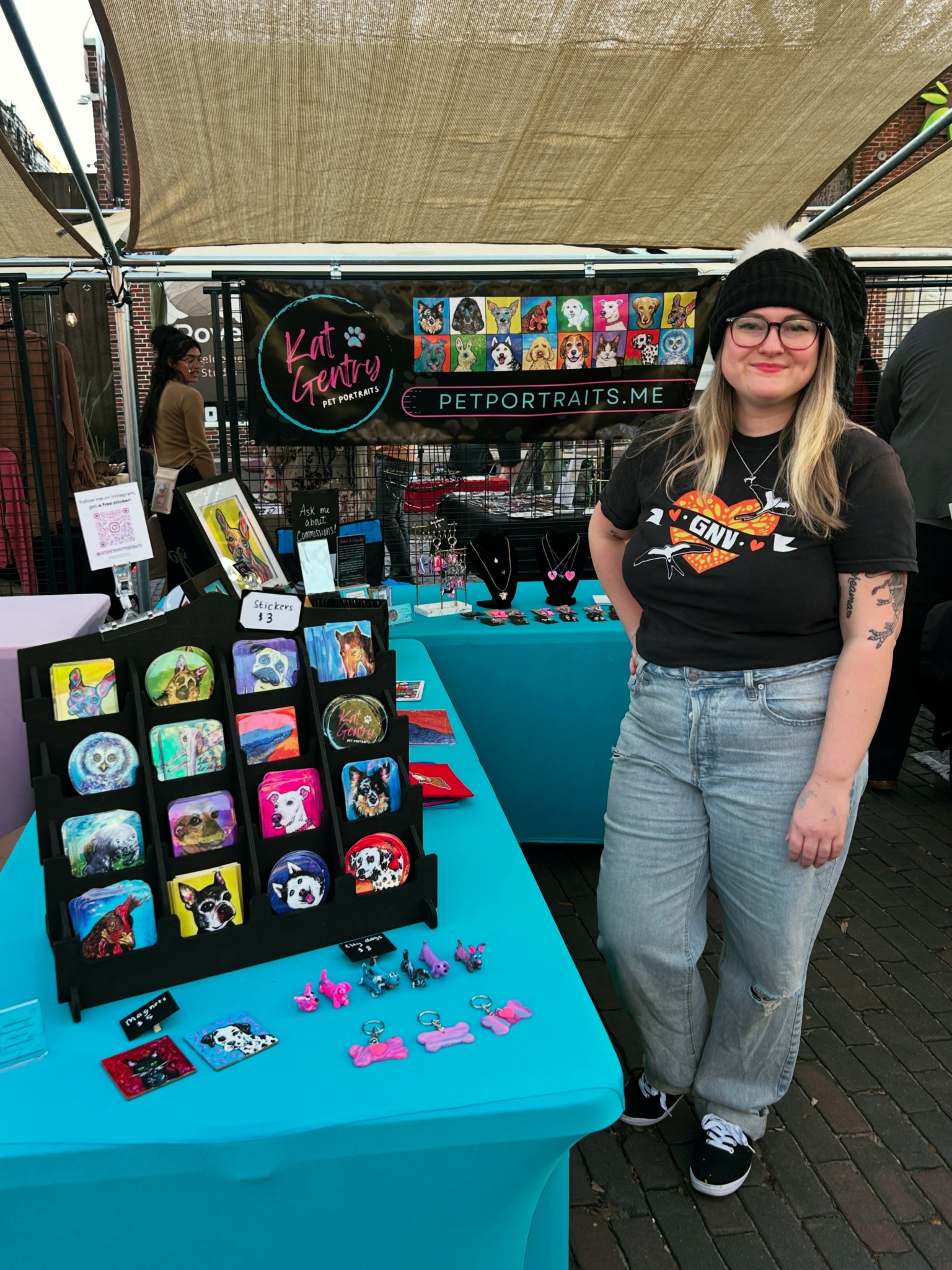 Woman with glasses and black beanie standing next to a booth with colorful artwork and pet portraits.